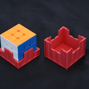 May include: A multi-colored 3x3 puzzle cube with orange, white, and blue faces, resting inside a red plastic square base. Beside it sits a matching red plastic square container with a castellated top edge, designed to hold the puzzle cube.