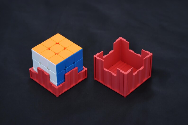 May include: A multi-colored 3x3 puzzle cube with orange, white, and blue faces, resting inside a red plastic square base. Beside it sits a matching red plastic square container with a castellated top edge, designed to hold the puzzle cube.