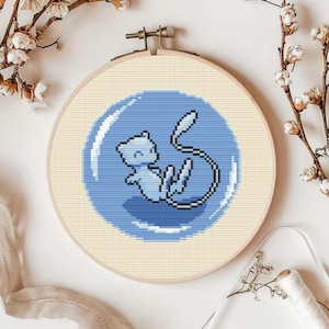May include: A cross-stitch embroidery hoop featuring a light blue cartoon character inside a blue bubble. The character has a long tail and is set against a cream-colored fabric. The wooden hoop is visible, and the background is white.