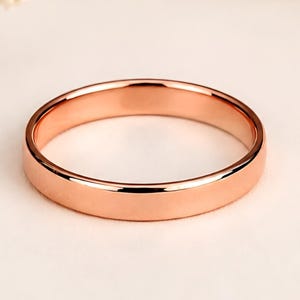 May include: A polished rose gold band ring with a smooth, flat surface and a rounded profile. The metal has a warm, metallic pink hue that reflects light evenly across its circular shape, showcasing a simple and clean design without any stones.