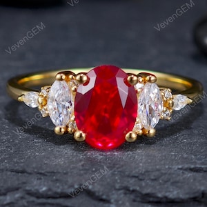 May include: A gold ring with a central oval-cut ruby, two marquise-cut white gemstones, and small diamonds. The ring is set on a delicate band. The background is dark, highlighting the ring's features.