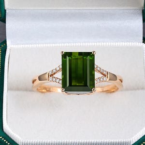 May include: A gold ring featuring a large, rectangular, emerald-cut green gemstone set in the center. The band is accented with small, clear, round-cut stones along the split shank, presented inside a dark green velvet jewelry box.