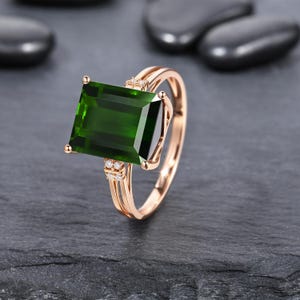 May include: A rose gold ring featuring a large, rectangular, emerald-green gemstone set in the center. The band is split into two thin, polished rose gold shanks, with small, clear, round accent stones embedded at the base of the main setting.