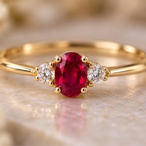 May include: A gold ring featuring a central oval-cut red ruby flanked by two small round-cut white diamonds. The stones are set in a polished yellow gold band with a thin, smooth profile, highlighting the contrast between the red and white gems.