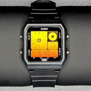 May include: A black digital watch with a rectangular face showing the time and date. The watch face has a yellow and orange display with the brand name "CASIO" at the top. The watch is on a black surface.
