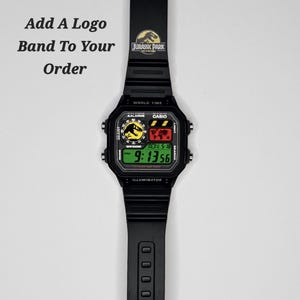May include: A black digital wristwatch with a rectangular face and a black rubber strap. The watch features a Jurassic Park logo on the band and display. The screen shows the time 9:13:56, a world map, and the text Add A Logo Band To Your Order.