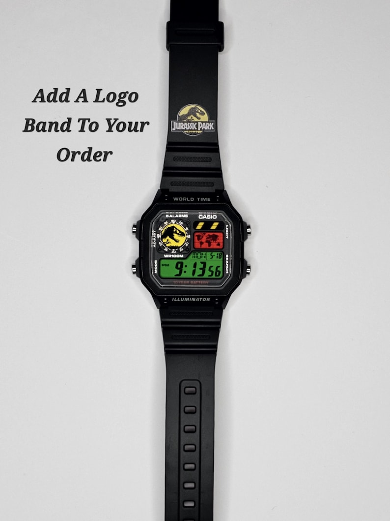 May include: A black digital wristwatch with a rectangular face and a black rubber strap. The watch features a Jurassic Park logo on the band and display. The screen shows the time 9:13:56, a world map, and the text Add A Logo Band To Your Order.