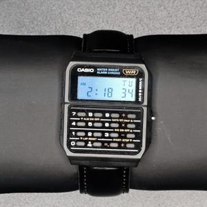 Może przedstawiać: Czarny cyfrowy zegarek Casio z klawiaturą kalkulatora i czarnym skórzanym paskiem. Tarcza zegarka wyświetla godzinę 2:18:34 AM. Zegarek jest wodoodporny i posiada funkcje alarmu i chronografu.