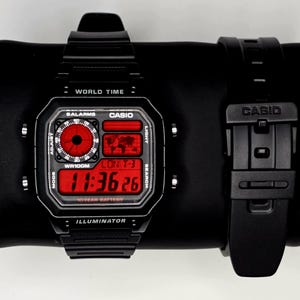Puede incluir: Reloj digital negro con una esfera rectangular y una correa negra a juego. La esfera del reloj muestra la hora en dígitos rojos, junto con otras funciones. El reloj tiene la palabra "CASIO" en la esfera y la correa.