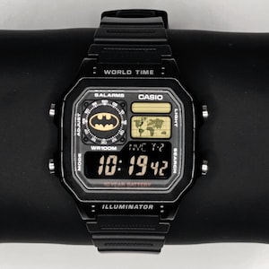 May include: Black Casio digital watch with a Batman logo on the face. The watch displays the time, date, and world time functions. The watch has a black band and a rectangular face with the text "WORLD TIME" and "ILLUMINATOR".