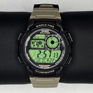 Puede incluir: Reloj digital Casio negro y marrón con pantalla digital verde. La esfera del reloj muestra la hora, la fecha y las funciones de hora mundial. El reloj tiene un bisel negro y una correa marrón. El reloj está etiquetado como "World Time" e "Illuminator".