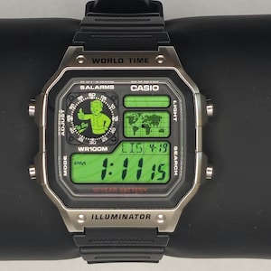 May include: A digital Casio World Time wristwatch with a black resin strap and a silver-tone bezel. The watch face features a green digital display, a world map graphic, and a circular Vault Boy character icon. Text includes WORLD TIME, CASIO, 5 ALARMS, WR100M, and ILLUMINATOR.