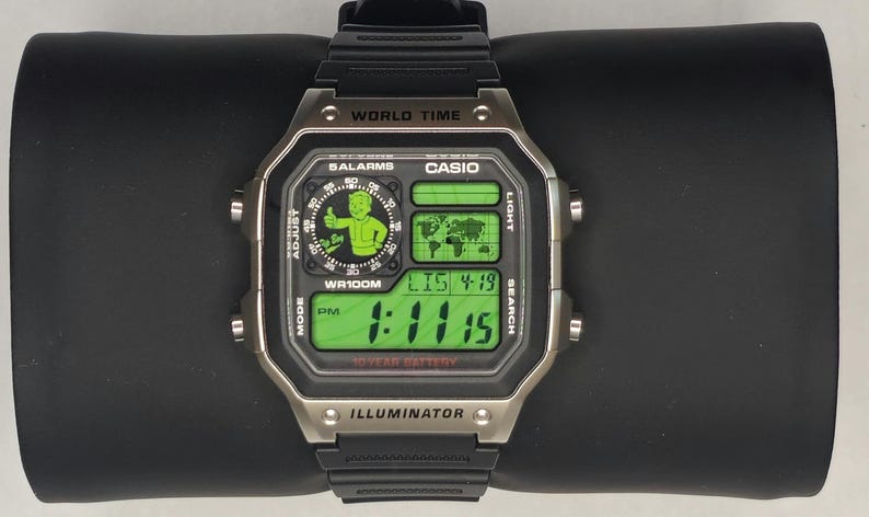 May include: A digital Casio World Time wristwatch with a black resin strap and a silver-tone bezel. The watch face features a green digital display, a world map graphic, and a circular Vault Boy character icon. Text includes WORLD TIME, CASIO, 5 ALARMS, WR100M, and ILLUMINATOR.