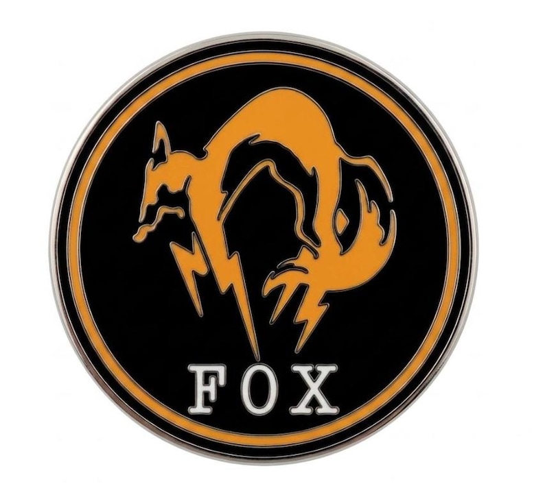 May include: A circular metal pin featuring a black background with an orange stylized fox silhouette and a lightning bolt design. The word FOX is printed in white capital letters at the bottom, all enclosed within a thin silver metallic border.