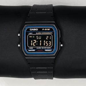 May include: Black Casio F-91W digital watch with a blue and black display. The watch shows the time as 12:11:53. The watch has a black band and is displayed on a black surface.
