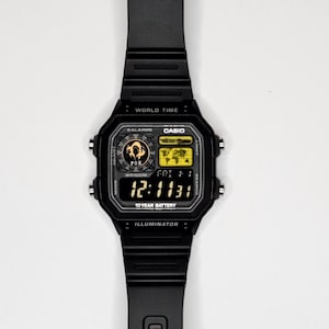 May include: A black digital wristwatch with a rectangular face, featuring a world map display, a fox graphic, and the text "WORLD TIME," "CASIO," "5 ALARMS," "WR100M," "10 YEAR BATTERY," and "ILLUMINATOR." The watch has a black resin strap and four side buttons.