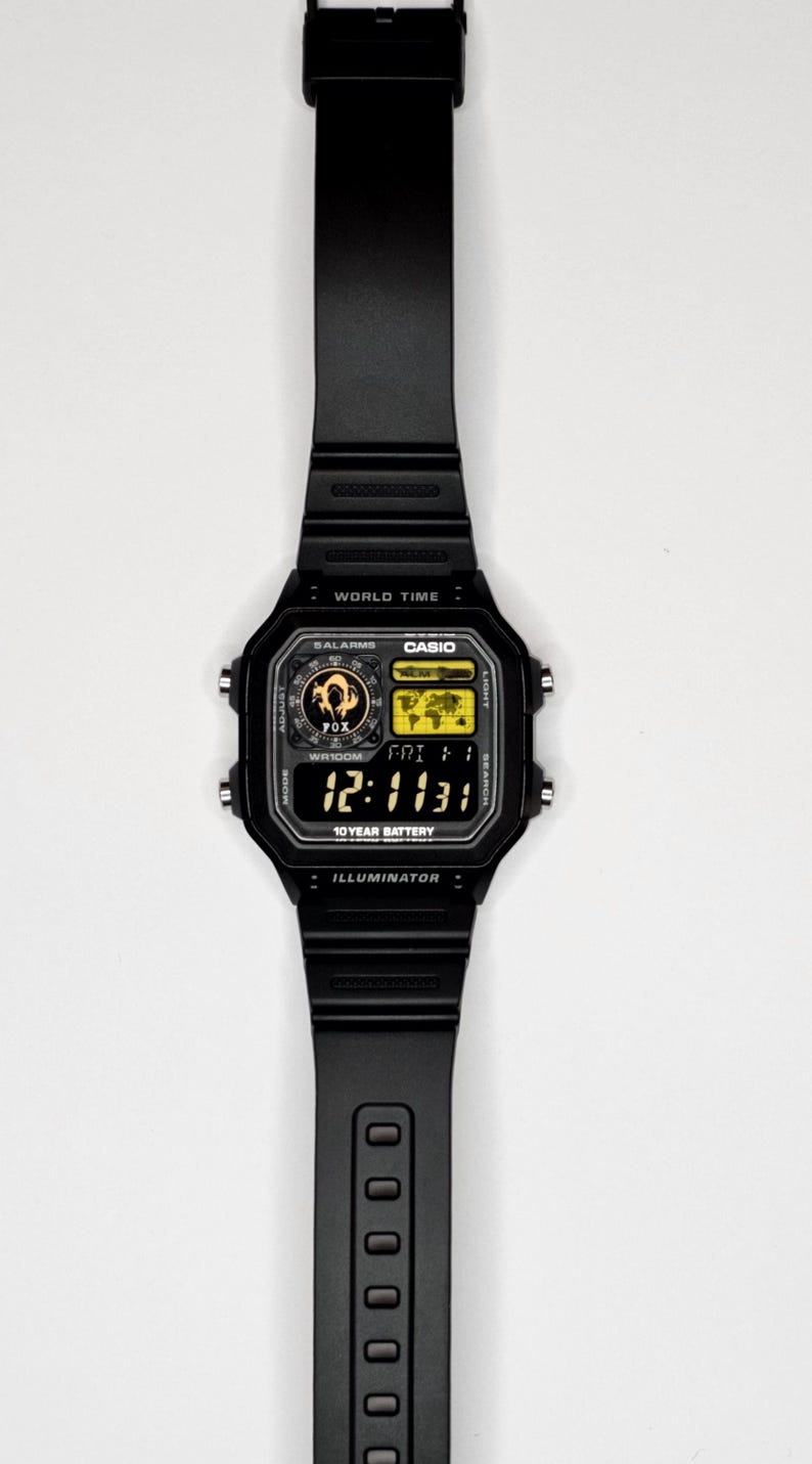 May include: A black digital wristwatch with a rectangular face, featuring a world map display, a fox graphic, and the text "WORLD TIME," "CASIO," "5 ALARMS," "WR100M," "10 YEAR BATTERY," and "ILLUMINATOR." The watch has a black resin strap and four side buttons.