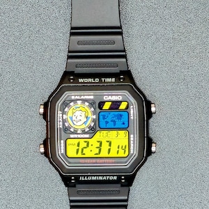Op de afbeelding: Zwarte Casio digitale horloge met een rechthoekige wijzerplaat. Het horloge geeft de tijd, datum en andere functies weer op een geel en blauw digitaal scherm. De horlogeband is zwart en gemaakt van flexibel materiaal. Het horloge heeft de tekst "WORLD TIME" en "ILLUMINATOR".
