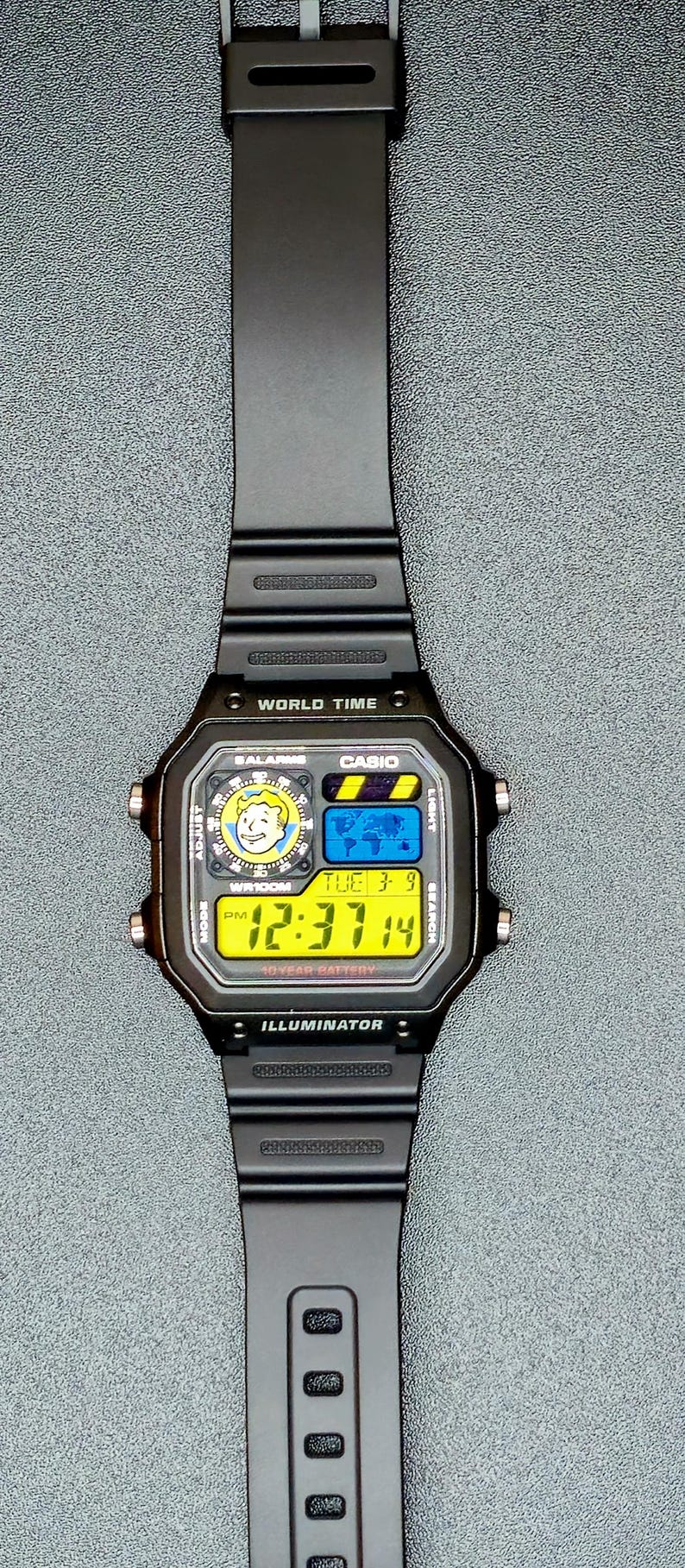 Op de afbeelding: Zwarte Casio digitale horloge met een rechthoekige wijzerplaat. Het horloge geeft de tijd, datum en andere functies weer op een geel en blauw digitaal scherm. De horlogeband is zwart en gemaakt van flexibel materiaal. Het horloge heeft de tekst "WORLD TIME" en "ILLUMINATOR".