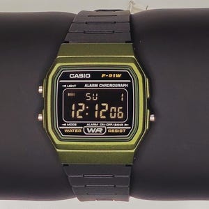 Może przedstawiać: Cyfrowy zegarek Casio F-91W z zieloną kopertą i czarnym paskiem. Tarcza zegarka pokazuje godzinę 12:12:06. Zegarek posiada logo Casio i tekst, w tym "ALARM CHRONOGRAPH" i "WATER RESIST".