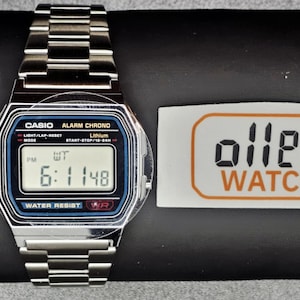 Puede incluir: Un reloj digital Casio plateado con una esfera negra y azul que muestra la hora 6:11 PM. El reloj está sobre una superficie negra junto a una pegatina que dice "ollee WATCH" en negro y naranja.