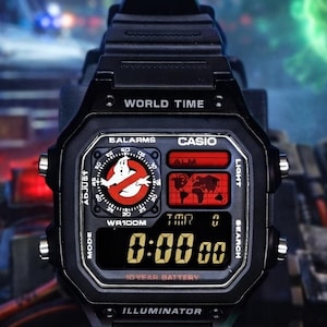 Könnte beinhalten: Schwarze Digitaluhr mit quadratischem Zifferblatt. Die Uhr zeigt das Ghostbusters-Logo, eine Weltkarte und den Text "WORLD TIME", "CASIO" und "10 YEAR BATTERY". Die angezeigte Uhrzeit ist 0:00:00.
