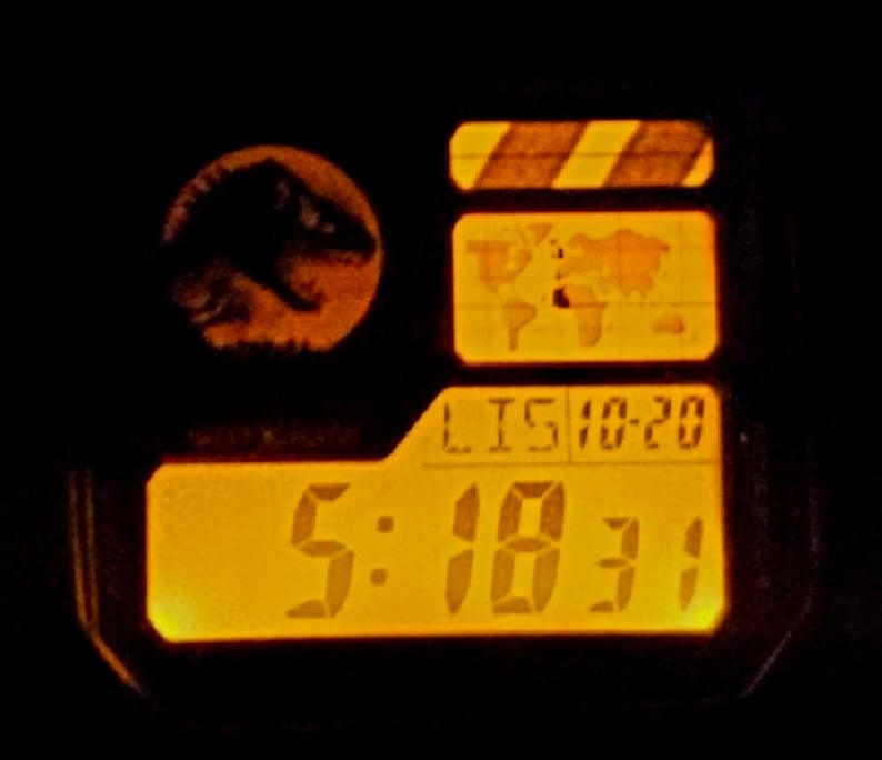 May include: A digital watch face with a bright orange backlight. The display features a circular dinosaur silhouette logo, a world map graphic, and the text LIS 10-20 above the digital time readout of 5:18:31. The watch has a dark frame.