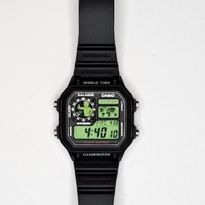 May include: A black digital wristwatch with a rectangular case and a resin strap. The watch face features a green backlit display showing the time 4:40, the date FRI 4-30, a world map, and a graphic of a cartoon character giving a thumbs up.