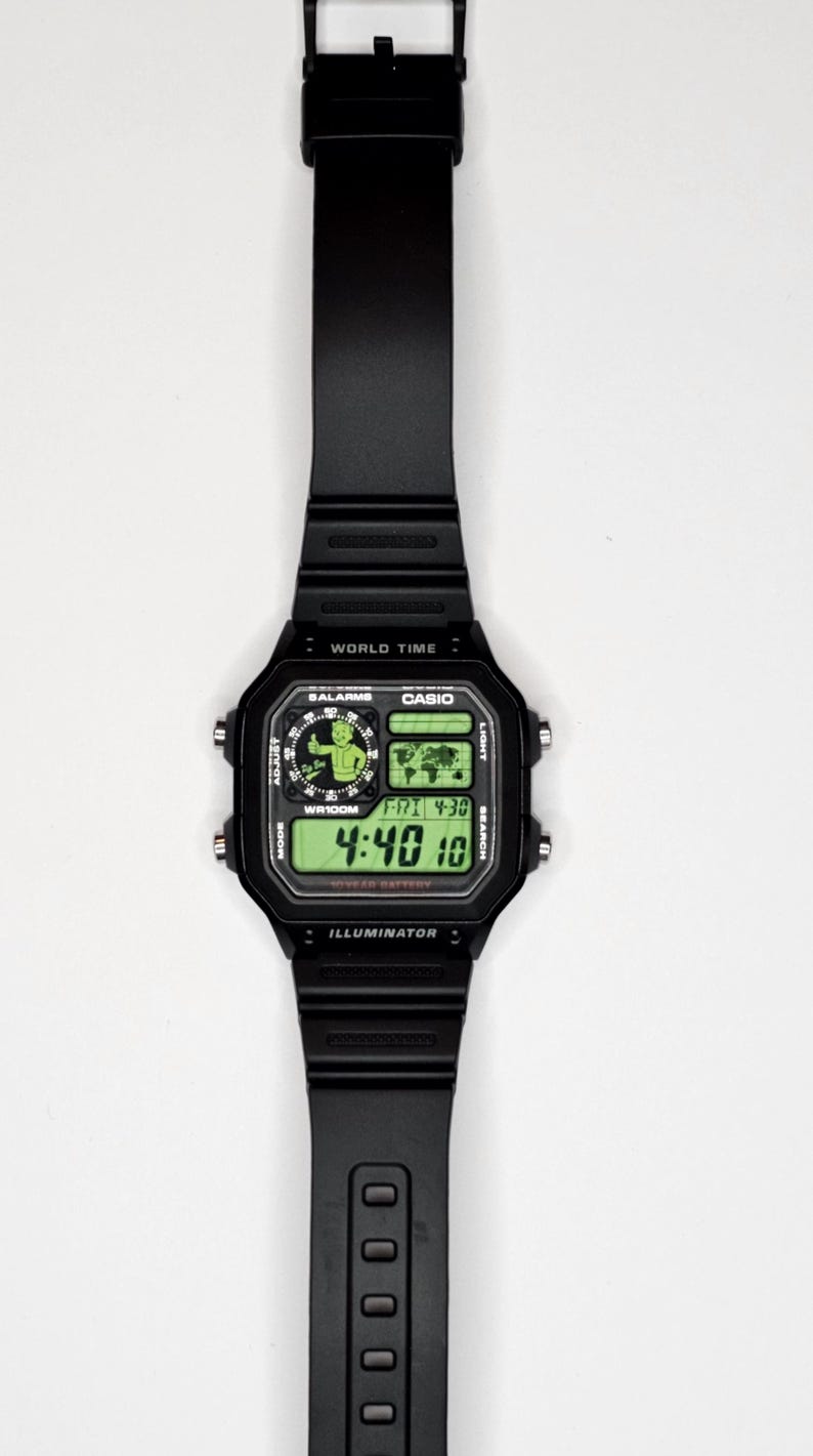 May include: A black digital wristwatch with a rectangular case and a resin strap. The watch face features a green backlit display showing the time 4:40, the date FRI 4-30, a world map, and a graphic of a cartoon character giving a thumbs up.