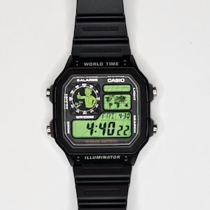 May include: A black digital wristwatch with a rectangular face, featuring a green backlit display. The screen shows a Fallout Vault Boy graphic, a world map, the text "WORLD TIME," "CASIO," "WR100M," "ILLUMINATOR," and the time 4:40:22.