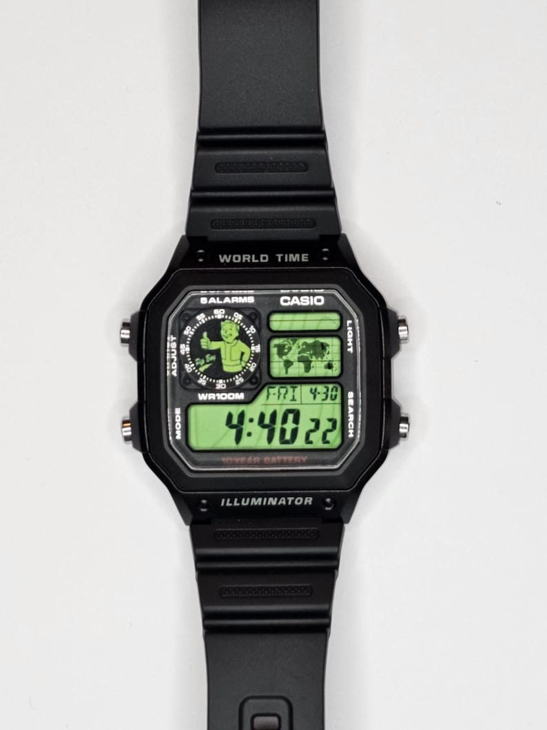 May include: A black digital wristwatch with a rectangular face, featuring a green backlit display. The screen shows a Fallout Vault Boy graphic, a world map, the text "WORLD TIME," "CASIO," "WR100M," "ILLUMINATOR," and the time 4:40:22.