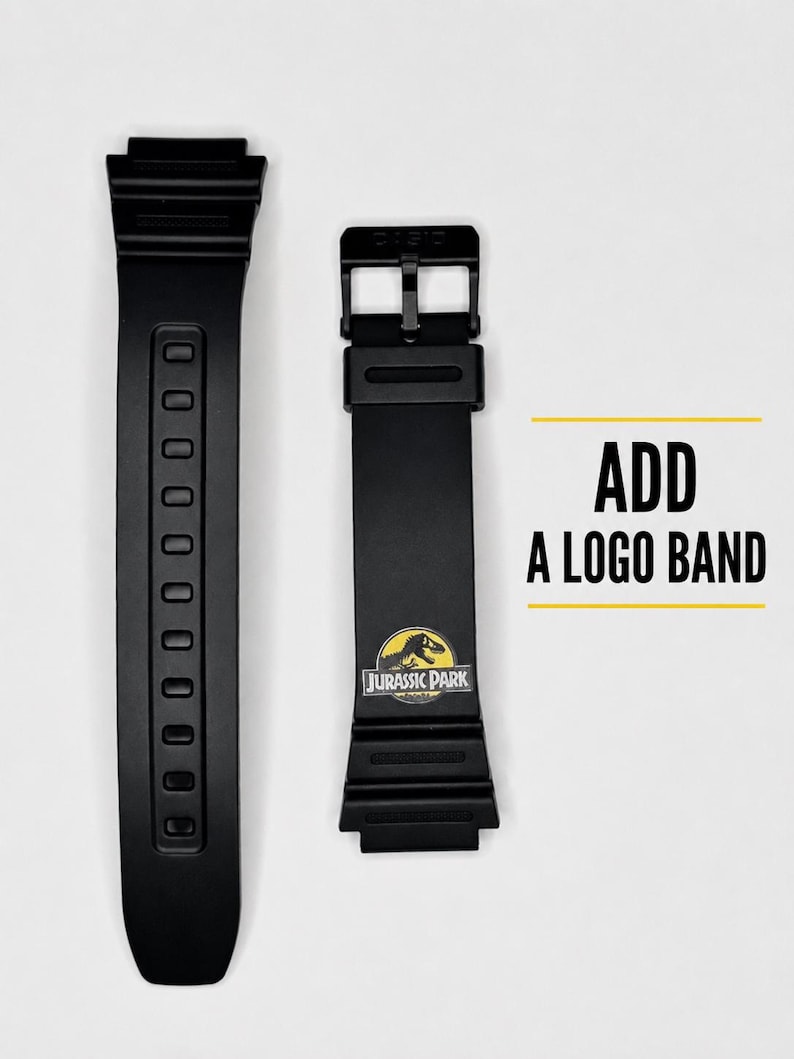 May include: Two black watch straps made of flexible material. One strap features a buckle and a yellow and black Jurassic Park logo graphic. The text ADD A LOGO BAND is printed in black next to the straps with yellow horizontal lines above and below.