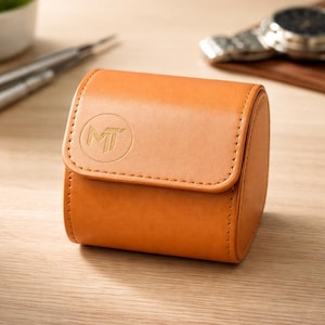 May include: A tan leather watch case with a flap closure. The case has a stitched border and a circular embossed logo on the front. The watch case is designed to protect and store a single watch.