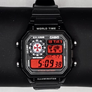 May include: Black Casio digital watch with a rectangular face displaying the time 5:09:38 in red digits. The watch has multiple functions, including world time and alarms. The watch is on a black surface.