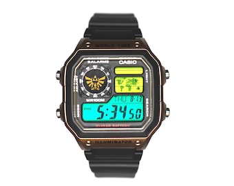 Könnte beinhalten: Eine digitale Casio-Armbanduhr mit schwarzem Kunststoffarmband und bronzefarbener Lünette. Das Display zeigt eine Weltkarte, ein Triforce-Symbol sowie den Text World Time, Illuminator und 10 Year Battery. Die Digitalanzeige zeigt 5:34:50 und das Datum.