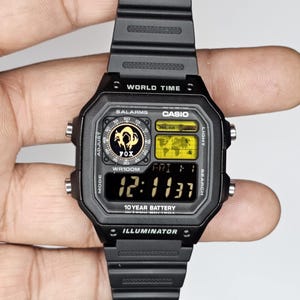 May include: A black Casio digital watch with a resin strap, featuring a square face with a world map display, a Fox logo, and the text World Time, 5 Alarms, WR100M, 10 Year Battery, and Illuminator. The digital screen shows the time 12:11:37 and date FRI 1.