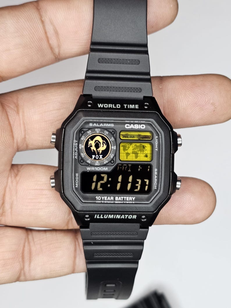 May include: A black Casio digital watch with a resin strap, featuring a square face with a world map display, a Fox logo, and the text World Time, 5 Alarms, WR100M, 10 Year Battery, and Illuminator. The digital screen shows the time 12:11:37 and date FRI 1.