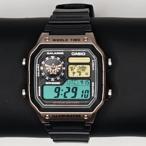 May include: A black and bronze Casio digital watch with a black band. The watch face displays the time, date, and world time features. The watch has the text "WORLD TIME", "CASIO", and "ILLUMINATOR".