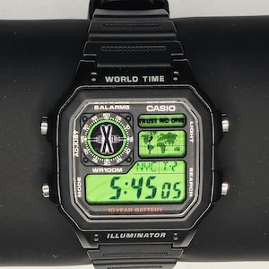 May include: Black Casio digital watch with a rectangular face displaying the time 5:45:05 in green digits. The watch has multiple functions, including world time and alarms. The watch is on a black surface.