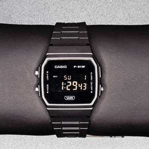Może przedstawiać: Czarny cyfrowy zegarek Casio F-91W z prostokątną tarczą i czarnym paskiem. Zegarek wyświetla godzinę, dzień i datę. Zegarek leży na czarnej powierzchni. Zegarek ma napis "CASIO", "F-91W" i "WR".
