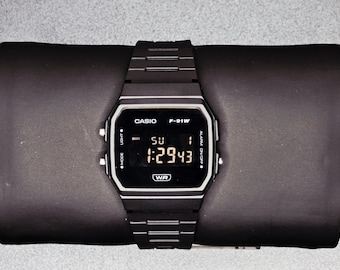 May include: A black Casio F-91W digital watch with a rectangular face and a black strap. The watch displays the time, day, and date. The watch is on a black surface. The watch has the text "CASIO", "F-91W", and "WR".