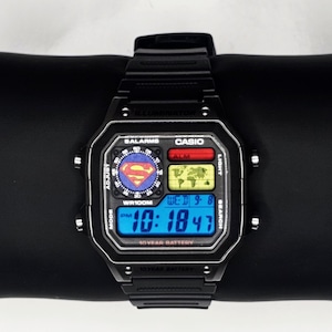 Puede incluir: Reloj digital Casio negro con el logotipo de Superman en la esfera. El reloj muestra la hora, la fecha y otras funciones en una pantalla digital azul. El reloj está sobre una superficie negra.