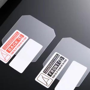 Puede incluir: Dos protectores de pantalla transparentes con pegatinas adjuntas. Una pegatina dice "BACK 1" y la otra "FRONT 2". Cada pegatina tiene instrucciones para quitar la m&aacute;scara. Los protectores est&aacute;n sobre una superficie oscura.