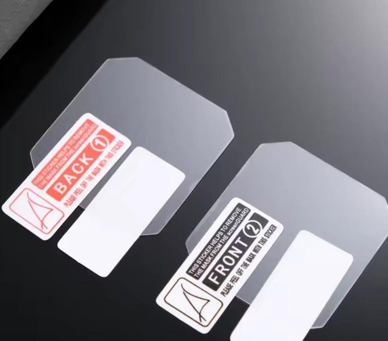 Puede incluir: Dos protectores de pantalla transparentes con pegatinas adjuntas. Una pegatina dice "BACK 1" y la otra "FRONT 2". Cada pegatina tiene instrucciones para quitar la m&aacute;scara. Los protectores est&aacute;n sobre una superficie oscura.