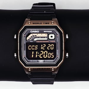 Op de afbeelding: Een zwart en goudkleurig digitaal Casio horloge met een zwarte band. De wijzerplaat toont de tijd en datum, samen met de tekst "WORLD TIME", "CASIO WR100M" en "ILLUMINATOR". Het horloge ligt op een zwarte ondergrond.