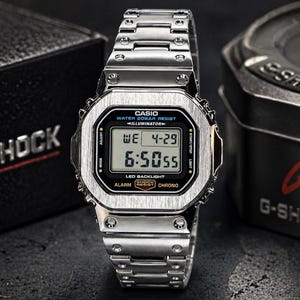 May include: A silver G-Shock digital watch with a rectangular face and a matching metal strap. The watch shows the date and time. The watch is next to a black box and a black tin, both with the G-Shock logo.