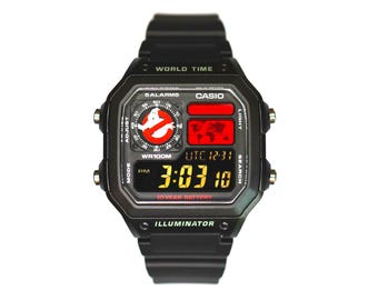 May include: A black Casio digital watch with a resin strap and square case. The display features a Ghostbusters logo, a world map, and the text 5 ALARMS, WORLD TIME, WR100M, 10 YEAR BATTERY, and ILLUMINATOR. The digital clock shows 3:03:10 PM.