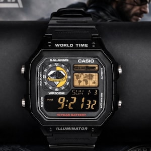 Puede incluir: Reloj digital Casio negro con esfera rectangular. El reloj muestra la hora, la fecha y las funciones de hora mundial. Incluye los textos "WORLD TIME", "5ALARMS" e "ILLUMINATOR". El reloj tiene una batería de 10 años.