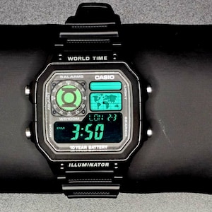 May include: Black Casio digital watch with a square face, displaying the time 3:50 in green digits. The watch features a world map and the text "ILLUMINATOR". The watch is on a black surface.