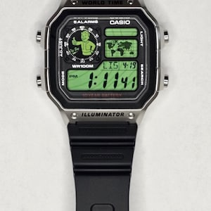May include: A black digital wristwatch with a silver-tone rectangular case and a black resin strap. The watch face features a green digital display with a world map, the text "WORLD TIME," "CASIO," "ILLUMINATOR," and a graphic of a cartoon character.